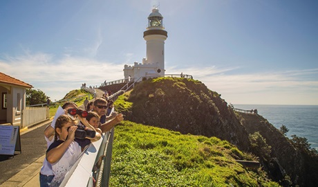 A group of people with binoculars watch for whales, with Cape Byron lighthouse in the background. Photo credit: John Spencer © DPIE