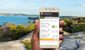 A hand holds a mobile phone with water views background from Sydney Harbour National Park. Photo: Simone Cottrell © DPIE